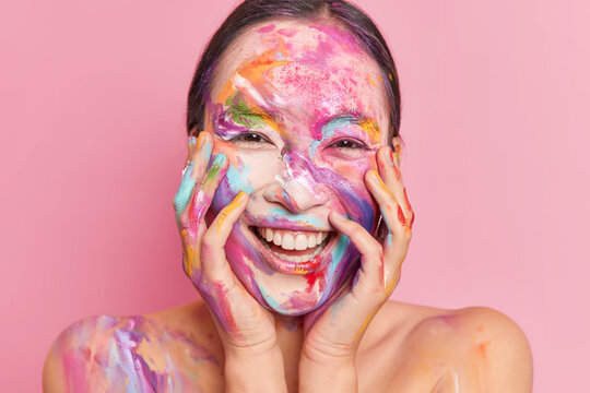 Headshot of happy brunette woman has face stained with colorful stains smiles broadly has white teeth keeps hands on cheeks isolated over rosy background. New creative makeup. Holi festival of colors