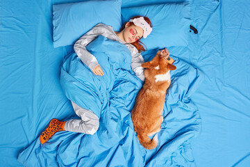 Young ginger woman resting on bed with dog sleeps deeply wears soft sleepmask pajama and socks...