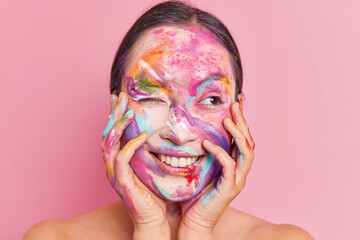 Positive young woman in colorful face paint has abstract makeup painting smears winks eyes smiles...