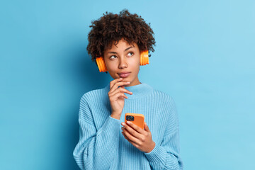 Photo of beautiful African American woman listens music with smartphone and headpones has thoughtful expression dressed in knitted jumpper isolated over blue background. Quarantine leisure concept © WHstudio Leushin N