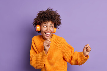 Happy curly haired fro American teenager dances carefree and sings favorite song listens music in headphones enjoys sound pleasant melody looks positively aside moves against purple background.