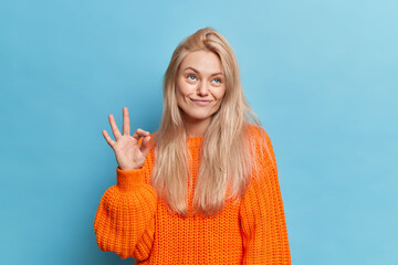Photo of pleased toughtful young woman with natural blonde hair makes okay gesture stabds pensive...