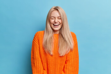 Overjoyed woman with blonde long hair laughs positively hears something funny closes eyes shows...