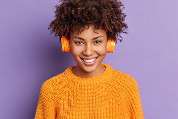 Close up shot of happy female music lover wears wireless stereo headphones on ears enjoys favorite song from playlist smiles positively poses against purple background. People hobby lifestyle concept