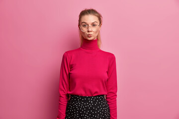 Good looking teenage girl folds lips as if wanting to kiss someone wears big transparent glasses...