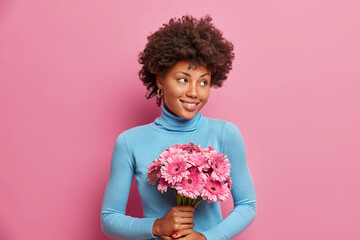 Pleased romantic African American woman holds bouquet of gerberas gets flowers during date with boyfriend smiles gently dressed in casual turtleneck isolated over pink background. Feminine concept