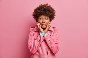Stressed worried African American woman bites finger nails looks nervously at camera afraids of bad consequences has curly bushy hair dressed in stylish jacket isolated over pink background.