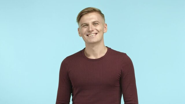 Attractive caucasian male model with blond hair looking at camera, start to laugh and smile with happy face, standing over blue background