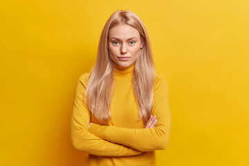 Obraz premium Studio shot of self confident blonde young woman keeps arms folded looks seriously at camera dressed in casual turtleneck isolated over yellow background. Pretty female model in assertive pose