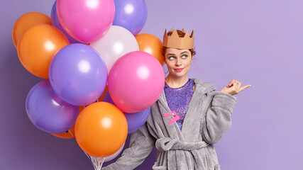 Fototapeta na wymiar Beautiful thoughtful woman in dressing gown crown points away on blank space holds big bunch of balloons going to congratulate someone with birthday shows direction to shop where to buy present