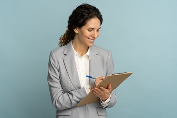Teacher woman wear grey blazer and white blouse writing, makes notes in document, holds folder in her hand. Female office worker isolated on studio blue background. 
