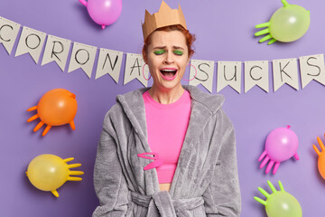Disappointed young woman wears crown and dressing gown expresses negative emotions cries indoor poses against purple background with balloons resembling virus upset because of coronavirus spread
