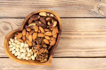 Natural nutritional blend of various nuts in a wooden plate in the shape of a heart symbol on a brown wooden table. A mixture of walnuts, brazil nuts, peanuts and almonds. Healthy natural food concept