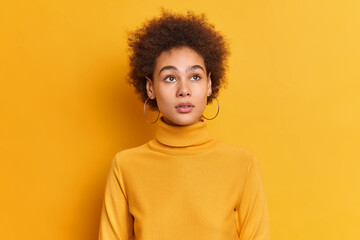 Fototapeta na wymiar Portrait of curly haired thoughtful woman concentrated somewhere has natural beauty wears casual turtleneck and earrings isolated over yellow studio wall. Pensive teenager thinks about future life