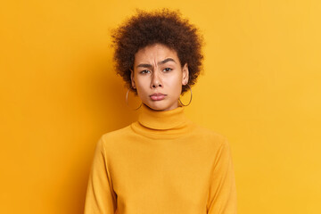 Fototapeta na wymiar Portrait of displeased millennial woman with Afro hair frowns face feels unhappy has some problems dressed in casual turtleneck isolated over yellow background. Negative emotions and feelings concept