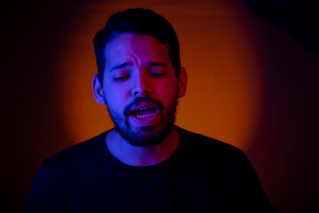 Fototapeta premium Young man portrait with colored lights in a studio shot with an orange light in the background. He is singing along with the music in his party.