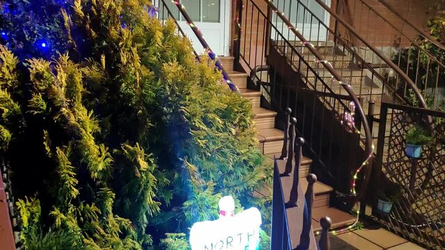 Entrance of a house decorated for Christmas. Wreath on the front door, a tree with blue flashing lights, a strip of ornamental lights around the handrail, and a bright light up sign saying "NorthPole"