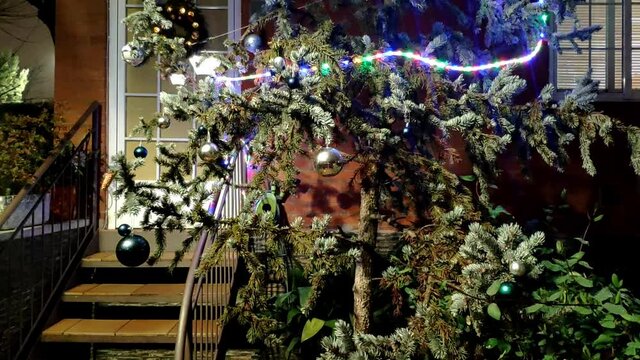 Big tree in front of a house entrance decorated for Christmas with big ornamental balls and a strip of ornamental coloured light with different colour dots going around it. The lights go on and off.