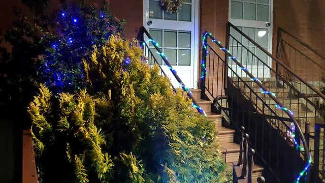 Entrance of a house decorated for Christmas. The front door has a wreath and there is a tree decorated with blue flashing lights. On the front steps handrail there is a ornamental lights strip.