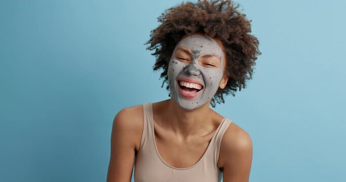 Happy dark skinned young woman raises palms and feels glad tries to frighten someone has fun applies facial clay mask dressed casually isolated over blue background. Beauty and skin care concept