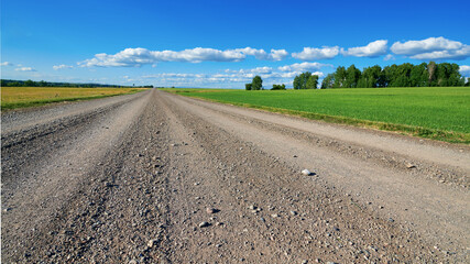 Gravel road close-up. Large stones are visible in the foreground. Around the green field and trees. Blue sky with few clouds in the distance. © Rauf