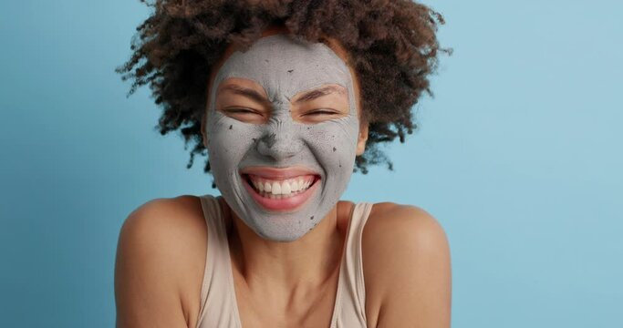 Optimistic funny curly woman closes eyes as awaits for something good happen smiles broadly looks at different sides applies clay mask undergoes skin care procedure isolated over blue background