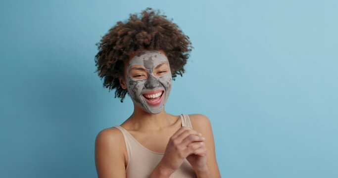 Beautiful overjoyed Afro American woman laughs positively smilesbroadly applies clay facial mask has skin care procedures after taking bath poses against blue background. Perfect well groomed face