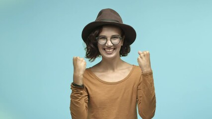 Beautiful girl in glasses and hipster hat, making fist pump with joy and screaming yes, celebrating victory, winning prize and looking happy, standing over blue background