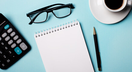 Glasses, calculator, blank notebook and pen on a light blue background. Copy space