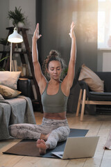 Fototapeta premium Young woman sitting on the floor in front of laptop and meditating online in the room