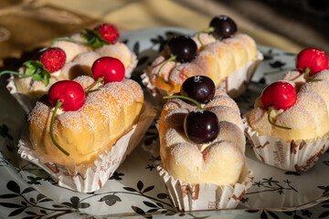 Artificial desserts presenting realistic sponge cakes coated with sugar with cherries and berries on top wrapped in baking paper, on a white glass tray with black ornaments during golden hour
