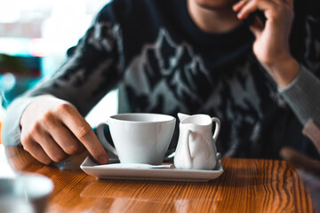 Man talking over the phone in a cafe with a coffee cup and cream milk pot on the table. Adult hand and winter sweater