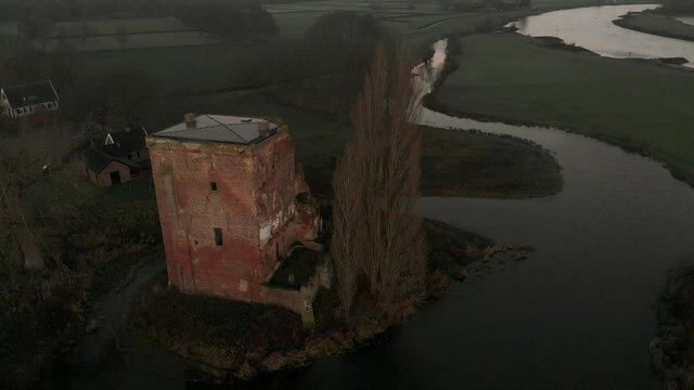 Slow aerial descend showing the remains of Nijenbeek castle revealing the early morning winter sunrise with ruins and tall tree in river delta landscape.