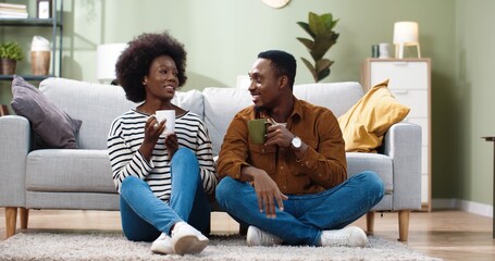 Nice lovely African American joyful married family couple man and woman sitting on floor, resting, drinking hot coffee or tea and speaking in good mood. Family leisure concept. Happy together