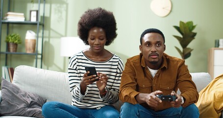 African American young handsome man sitting on sofa at home playing video games on console having fun while his wife typing and browsing online on mobile phone and showing him something Family concept