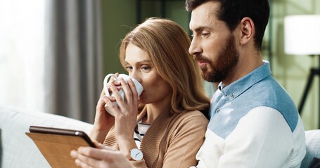 Portrait of young Caucasian joyful couple shopping on internet browsing online on tablet device sitting on sofa in apartment spending time together with cup of tea. Husband and wife resting at home