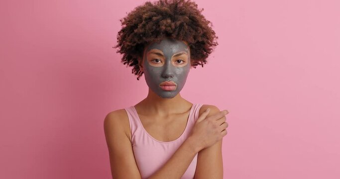 Young serious young woman with clay beauty mask on face to improve skin condition touches shoulder looks at camera has curly hair poses against rosy background. Pampering and wellness concept