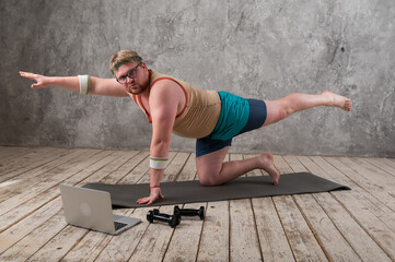 funny fat man in sports clothes doing exercises. He plays sports remotely while looking at his laptop