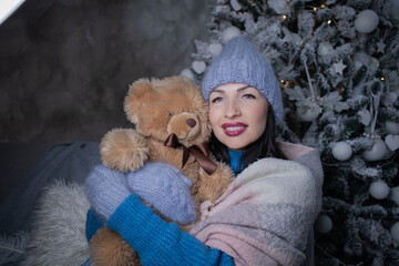 Fototapeta premium brunette in a blue sweater and blue hat and mittens with a teddy bear near the new year tree