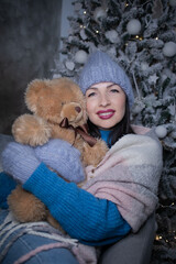 Fototapeta premium brunette in a blue sweater and blue hat and mittens with a teddy bear near the new year tree