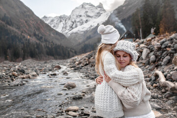 Fototapeta na wymiar Mother and daughter hug against the background mountain river, alpine view, snow on hills. Woman and girl dressed scandinavian style clothes, warm knitted hat, sweater.