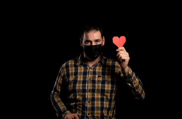 Closed portrait of cheerful young man holding a red paper heart in hand, isolated studio black background. Human facial expressions, emotions, feelings, body language, perception of life. Copy space