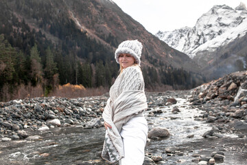 Young woman dressed in knitted warm hat, sweater and wrapping plaid blanket standing by the river and looking away to the adventure. Happy hiker wanderlust the view of snow, mountains