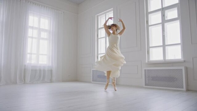 Ballerina spinning on tiptoe in pointe shoes in white fluttering dress in bright spacious studio