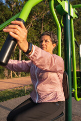 Elderly brown woman with pink jacket stretching arms in gym equipment, using smartwatch. Outdoors in sunny day. workout, effort, active, hard work, wellness concept.