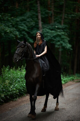 Young woman in hat and black cloak with a horse in the forest