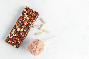 Scoop of whey or soy protein isolate, chocolate protein bar, white capsules of amino acids and creatine, bodybuilding food supplements on white background, top view, flat lay