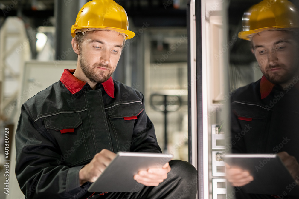Poster Young serious engineer in workwear and hardhat scrolling through technical data