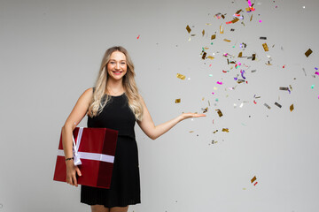 Smiling young girl with large festive gift pointing by hand on empty space with holiday confetti on gray background. High quality photo