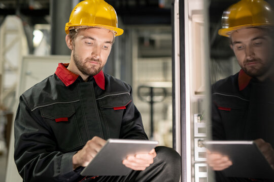 Young Serious Engineer In Workwear And Hardhat Scrolling Through Technical Data